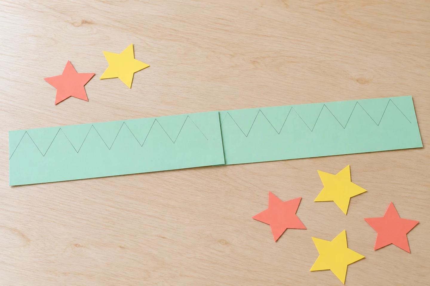 Two completely FLAT undecorated mint paper strips overlapping end to end, pencil guidelines for triangle tips, loose coral and yellow paper stars beside them