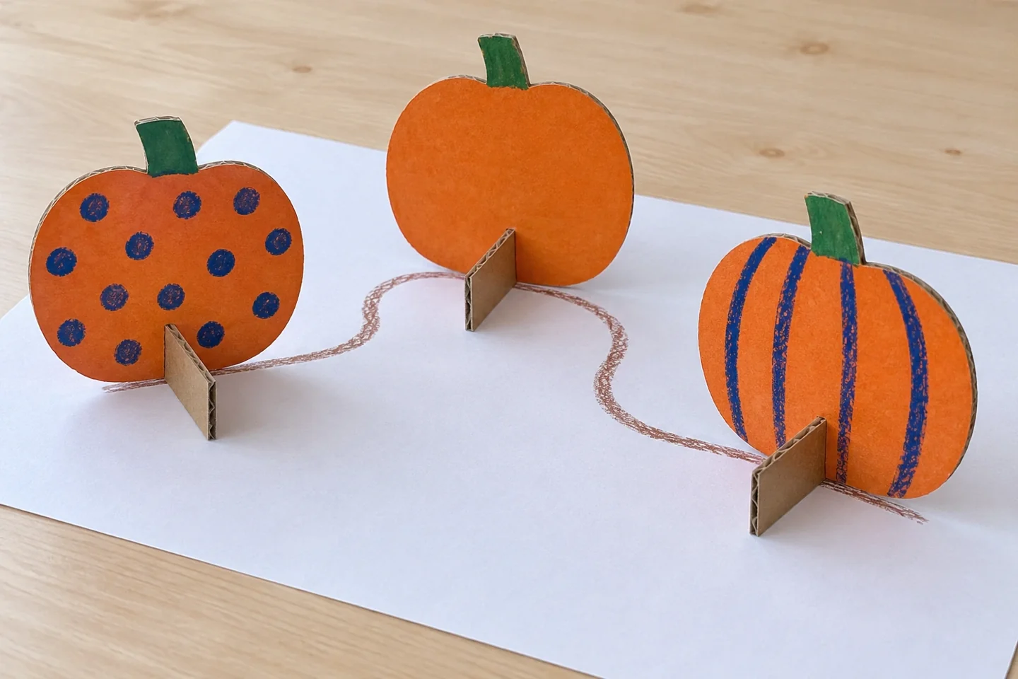 Three decorated cardboard pumpkins stand on perpendicular rectangular feet along a simple paper path.