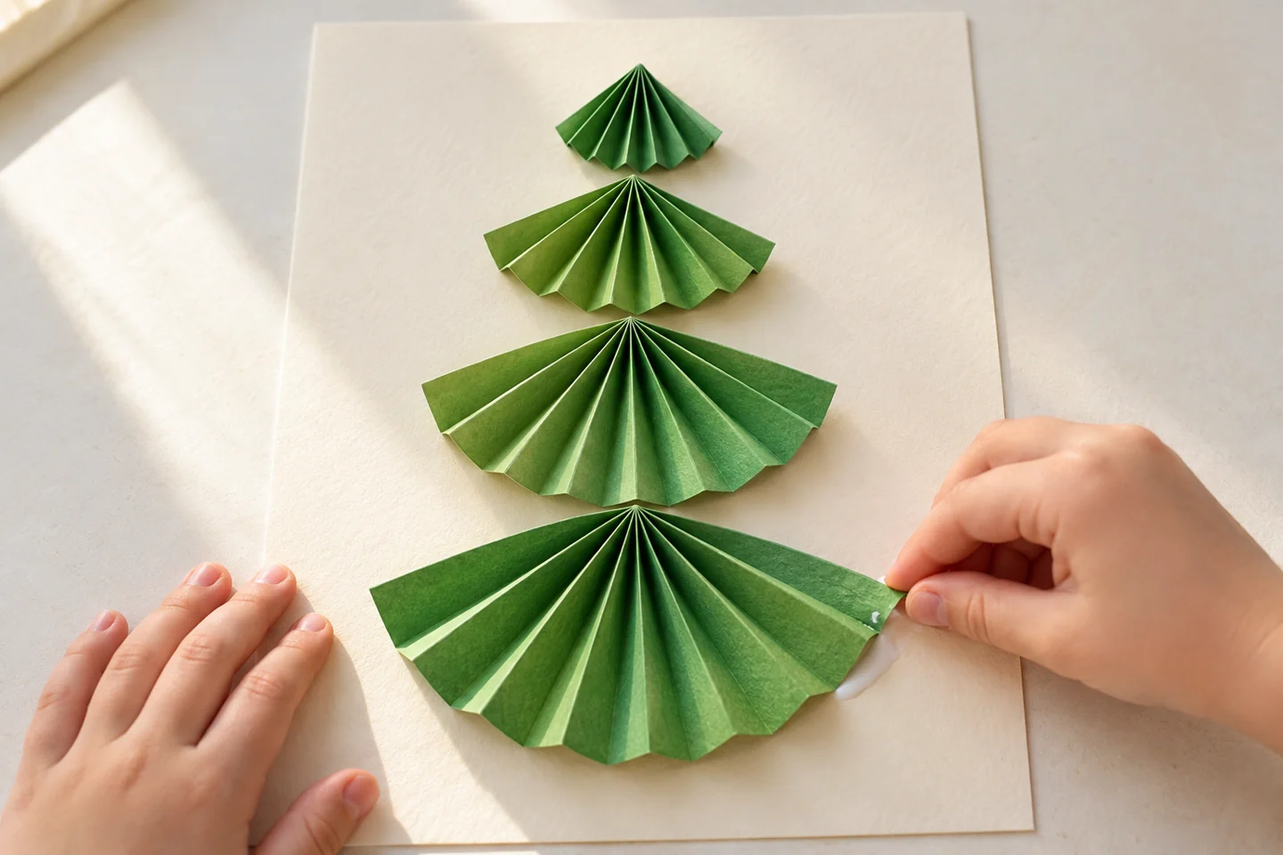 Close-up of four green accordion fans arranged on cream card from smallest at top to largest at bottom, pinched ends up, one fan edge being pressed onto glue.