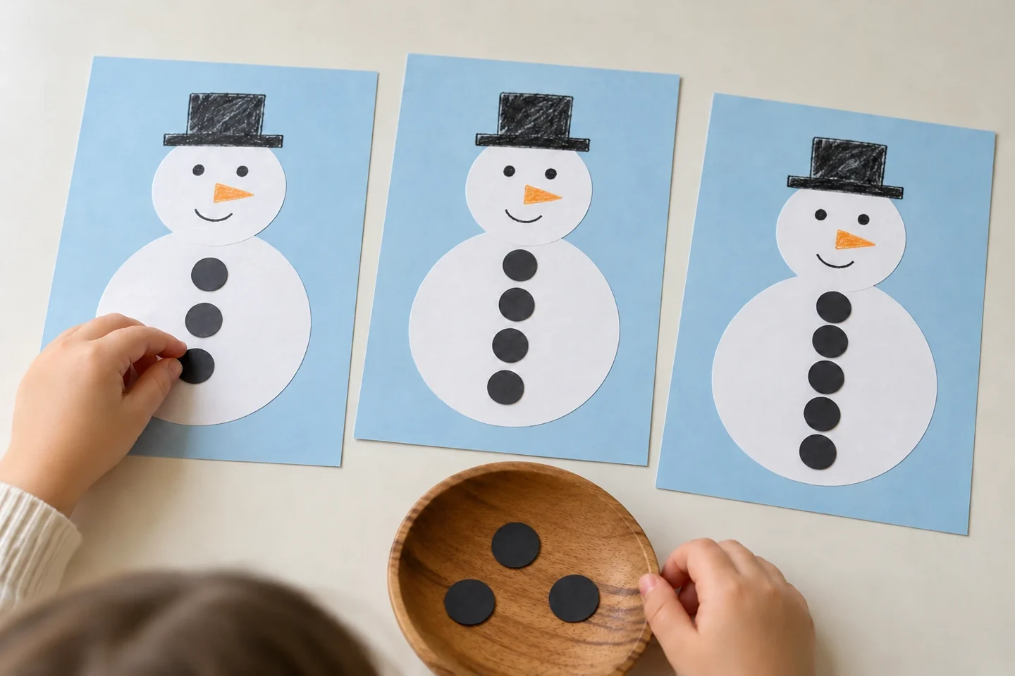 Child hands playing with three pale blue snowman counting mats and tray of dark paper circles